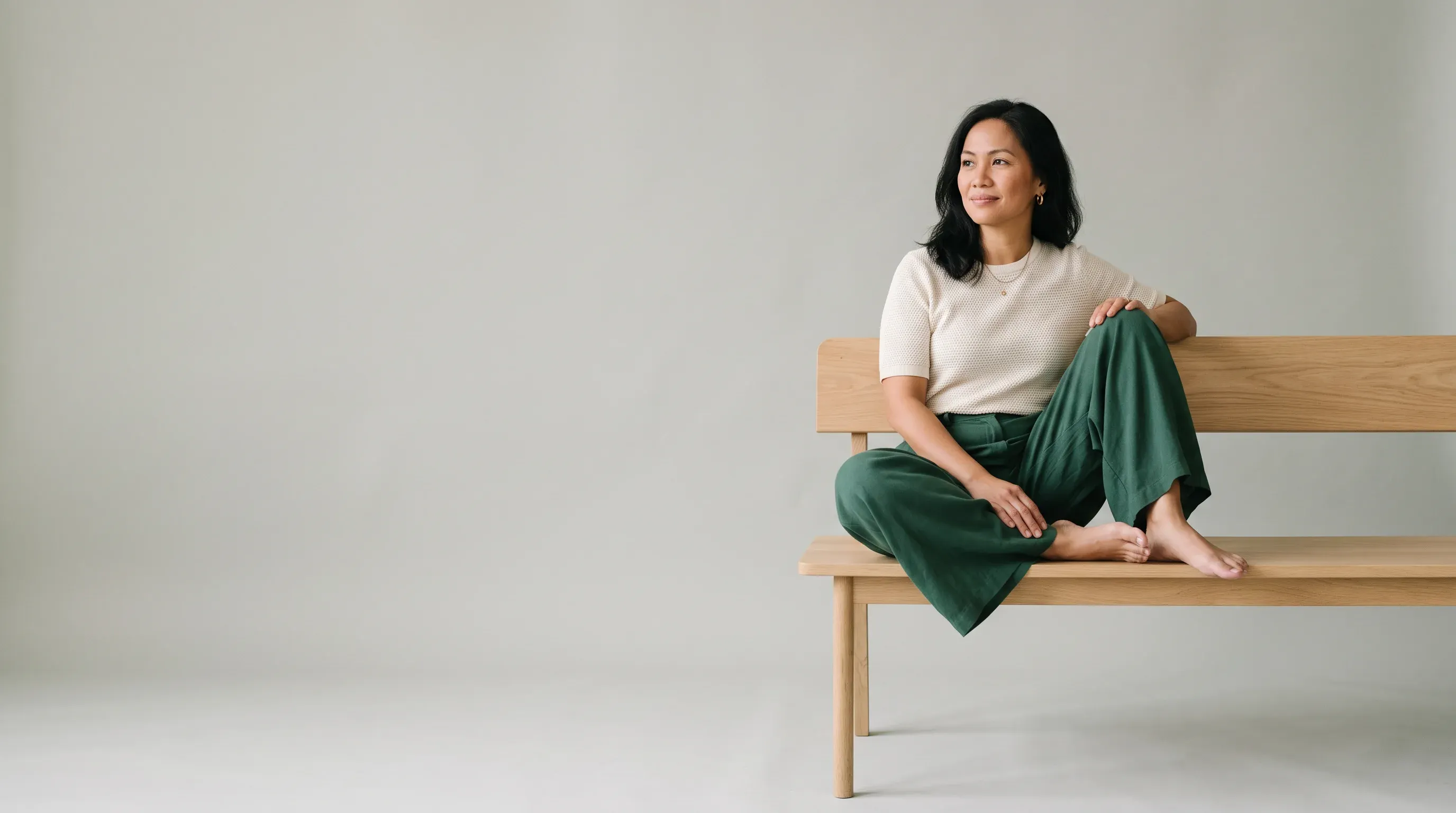 A woman in a cream top and green pants sitting on a wooden bench in a pale studio